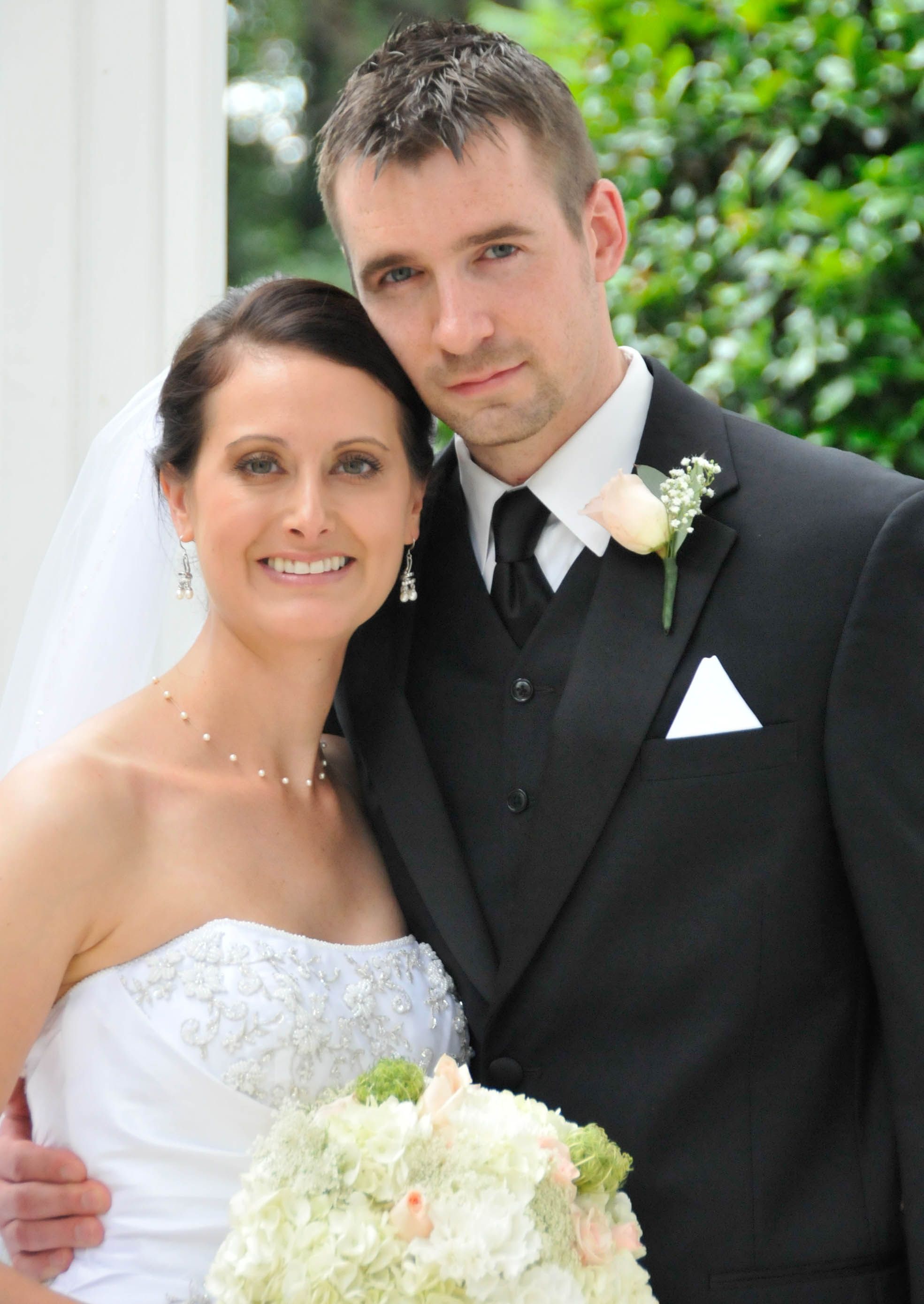 Wedding couple in formal attire posing with white floral bouquet in garden setting.
