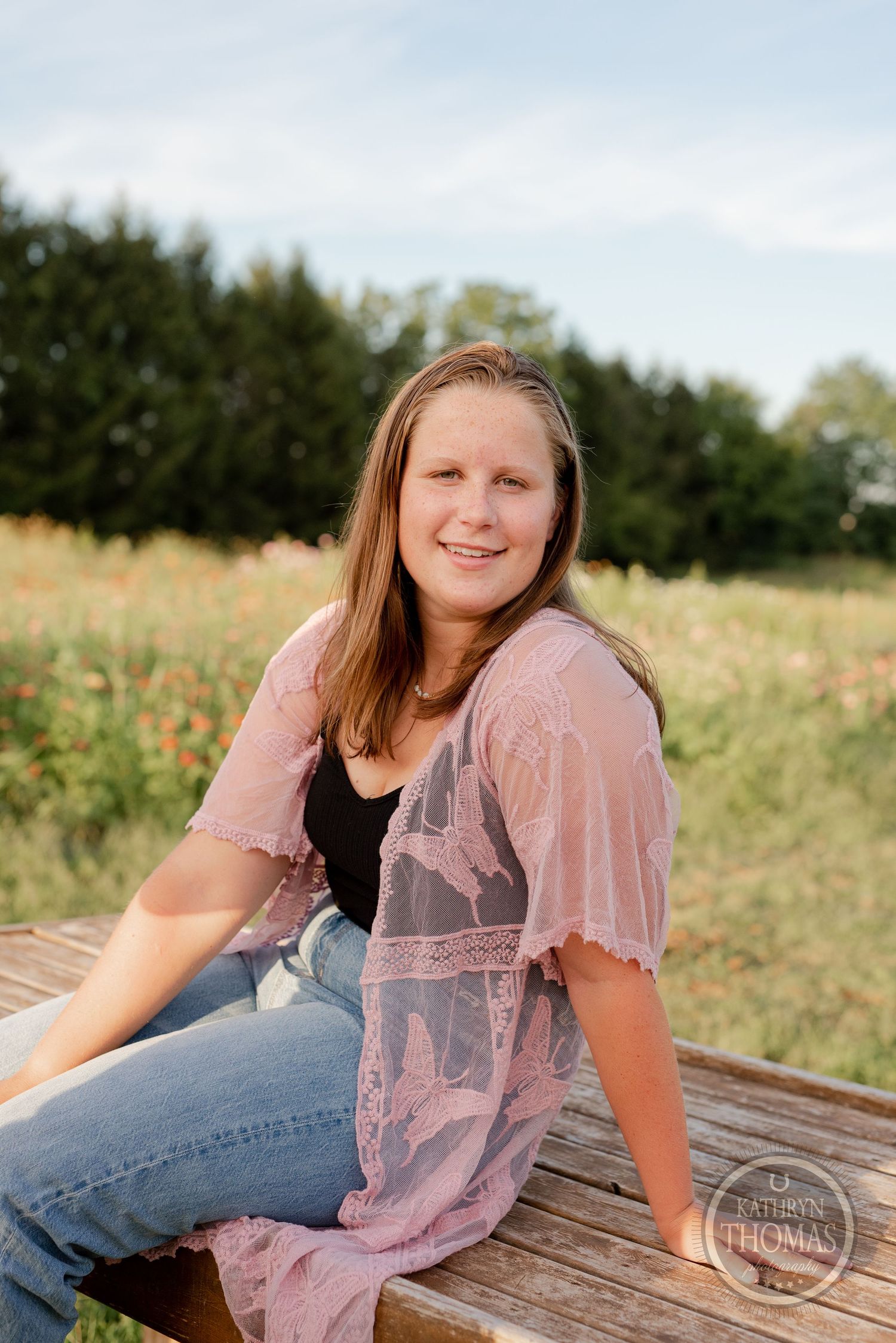Someone in pink lace and jeans sits on wooden surface in a summer field.