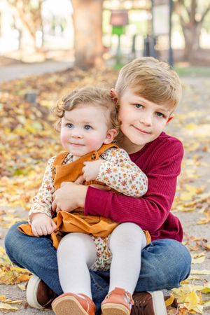Two children in autumn clothing sit together on fallen leaves during outdoor photo session.