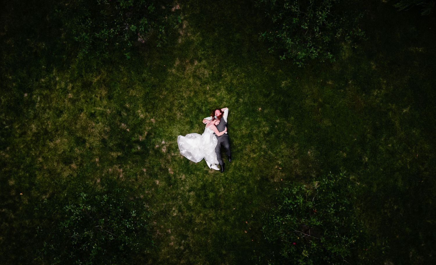 Aerial view of a couple in wedding attire lying on green grass, captured from above.