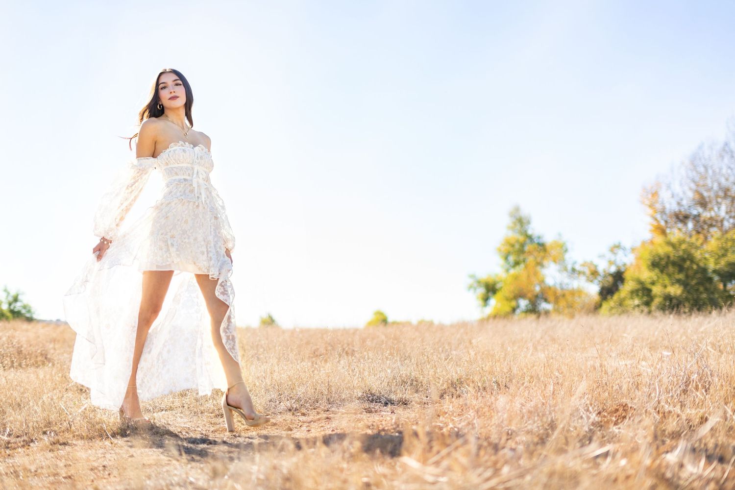 A high school senior in flowy white dress walking through golden field in sunlight.