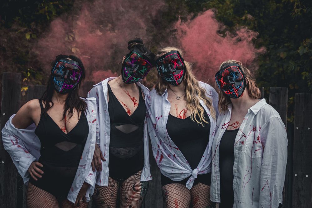 Four women in Halloween masks and zombie costumes pose with pink smoke bombs outdoors.