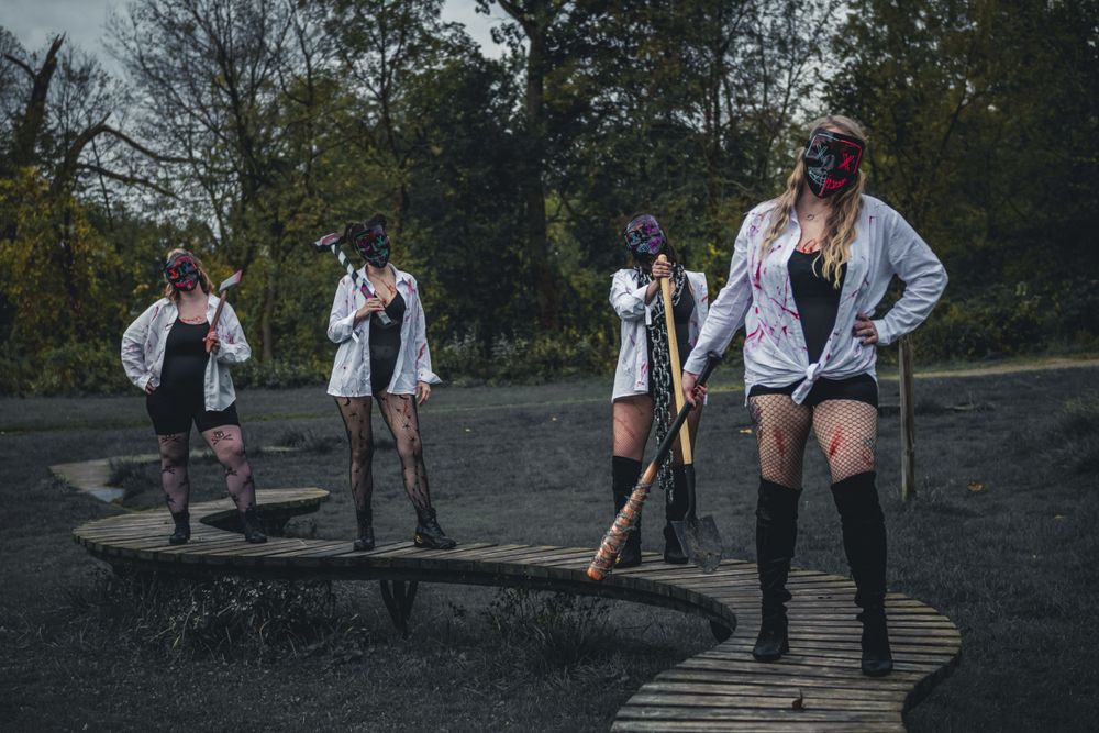 Four women in zombie costumes with masks holding weapons on a dark outdoor pathway.