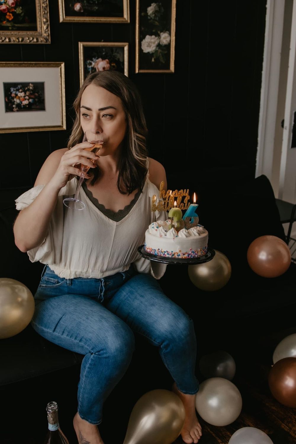 Woman holding birthday cake with candles while drinking champagne surrounded by gold balloons.