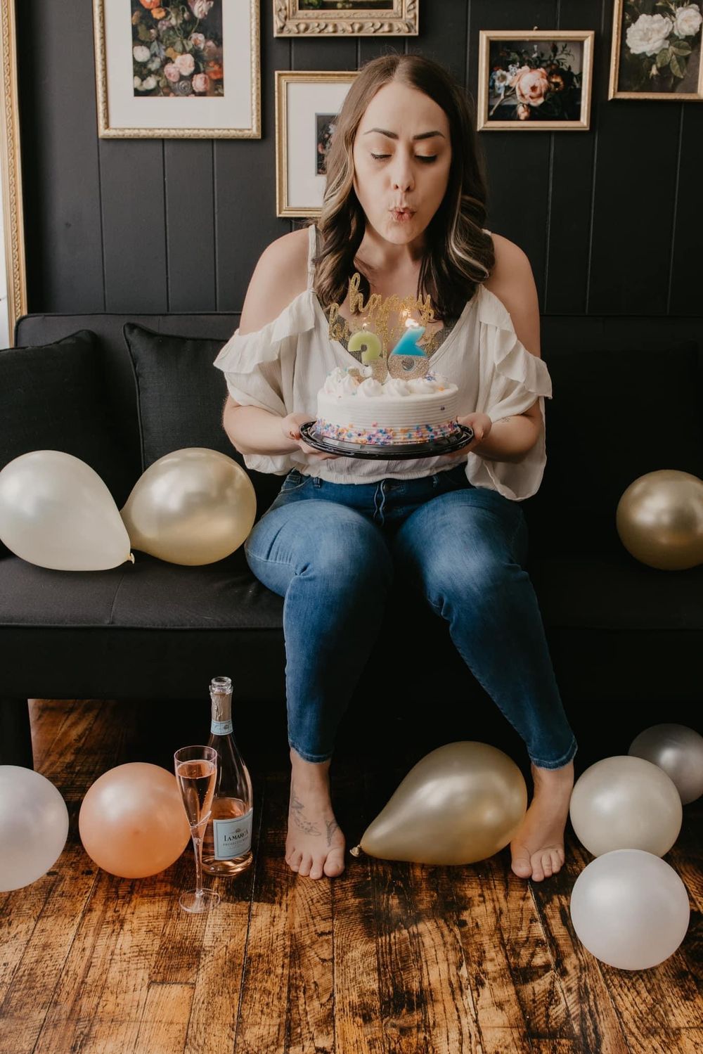 Woman blowing out birthday cake candles surrounded by balloons on dark sofa celebration.