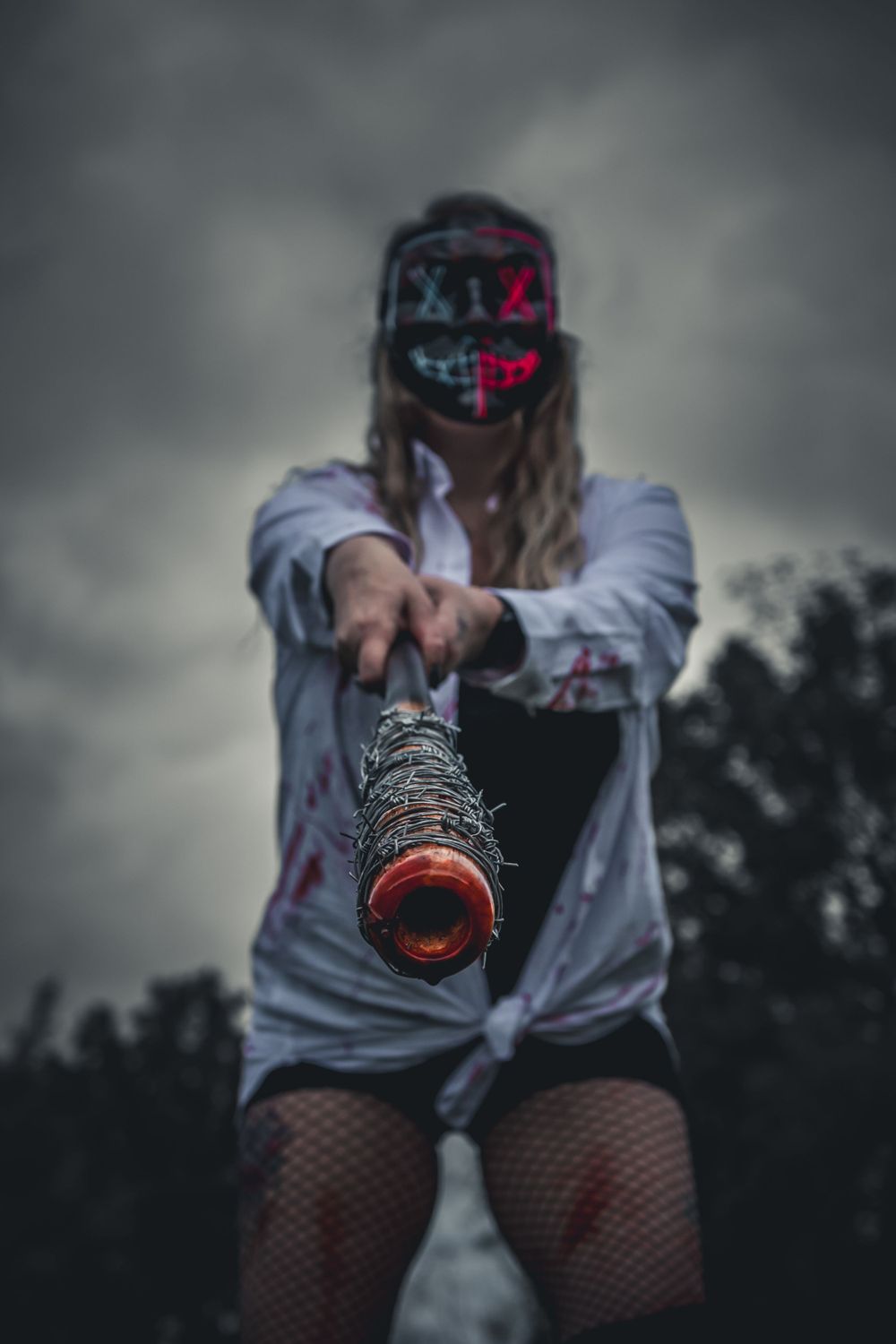 Woman in zombie costume holding barbed wire bat toward camera under dramatic cloudy sky.