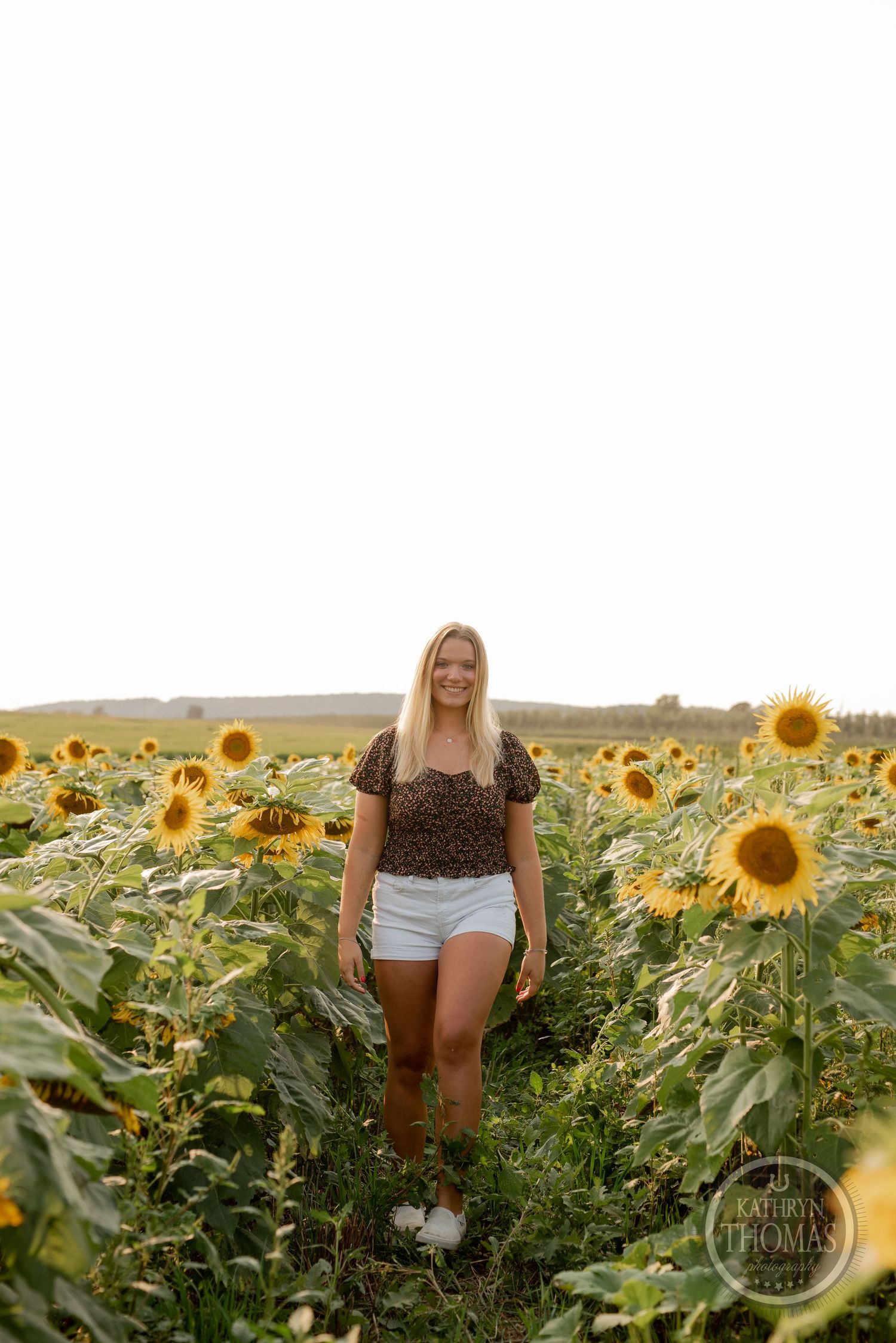 A full-length portrait of someone standing in a path between rows of towering sunflowers.