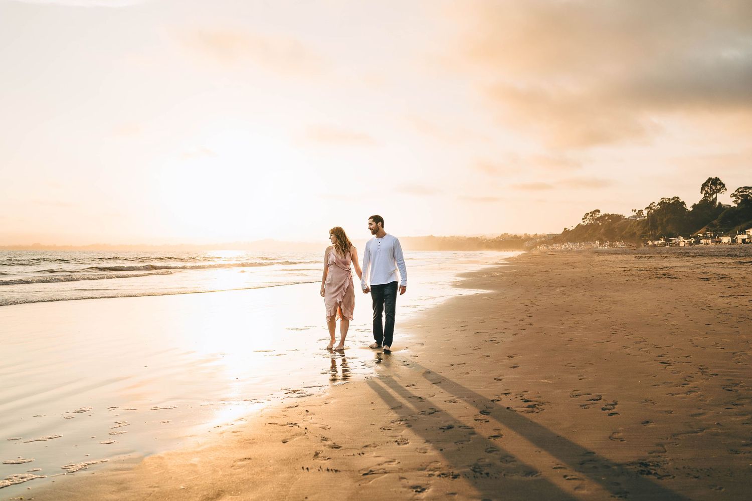 A couple walks hand in hand along a sandy beach during a golden sunset, casting long shadows on the shore.