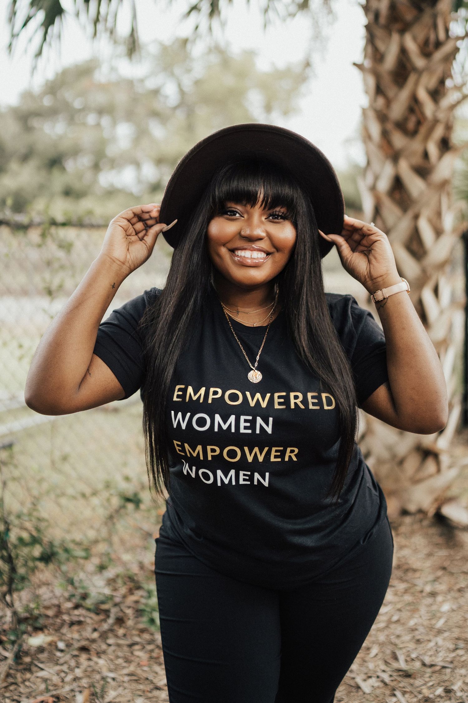 A person wearing a black t-shirt with empowering text smiles while adjusting their hat outdoors.