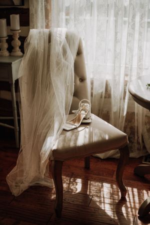 Elegant wedding veil and shoes artfully arranged on vintage chair by window.