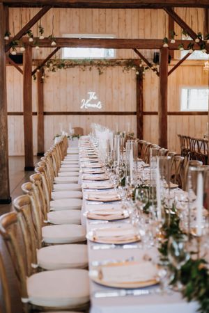 Long elegant wedding reception table with place settings and greenery in a rustic wood barn venue.