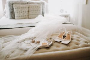 Wedding veil and white heeled sandals arranged on a neutral toned bed.
