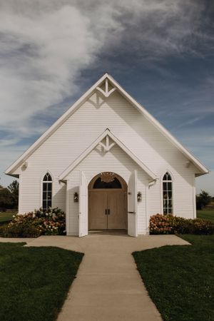 A charming white chapel with arched double doors and ornate architectural details stands against a dramatic cloudy sky.