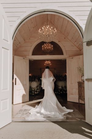 Dramatic silhouette of wedding gown under crystal chandeliers in white chapel doorway.