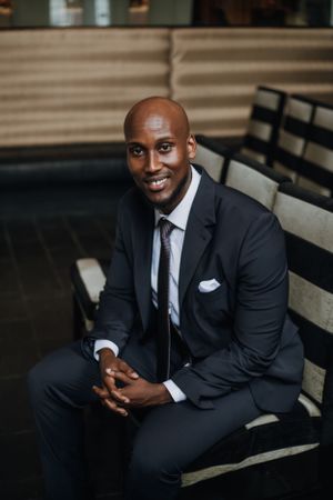 An elegantly dressed man in a dark suit sitting on a bench with a warm smile.