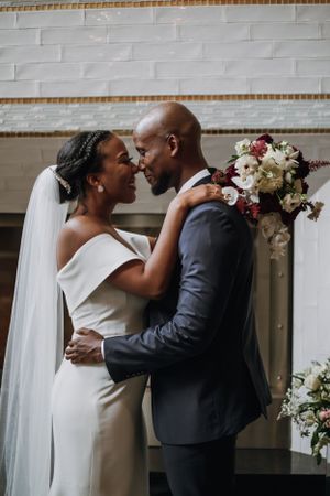 A romantic wedding portrait of newlyweds embracing near a decorated wall with floral arrangements.