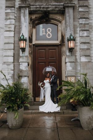 A couple shares an umbrella at a grand entrance with number 18 and lanterns.