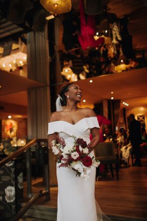 Elegant bride in off-shoulder white gown holding burgundy and white bouquet in candlelit venue.