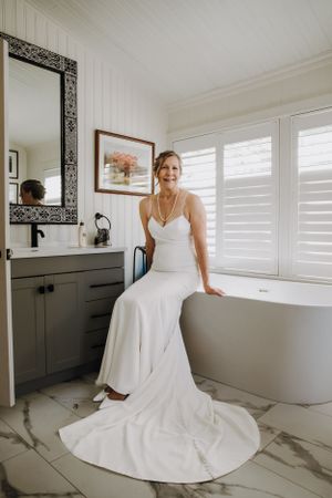 A bride in an elegant white gown poses in a modern bathroom with marble floors and shuttered windows.