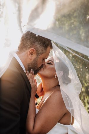 Romantic wedding couple sharing intimate moment under flowing veil with sunlight peeking through.