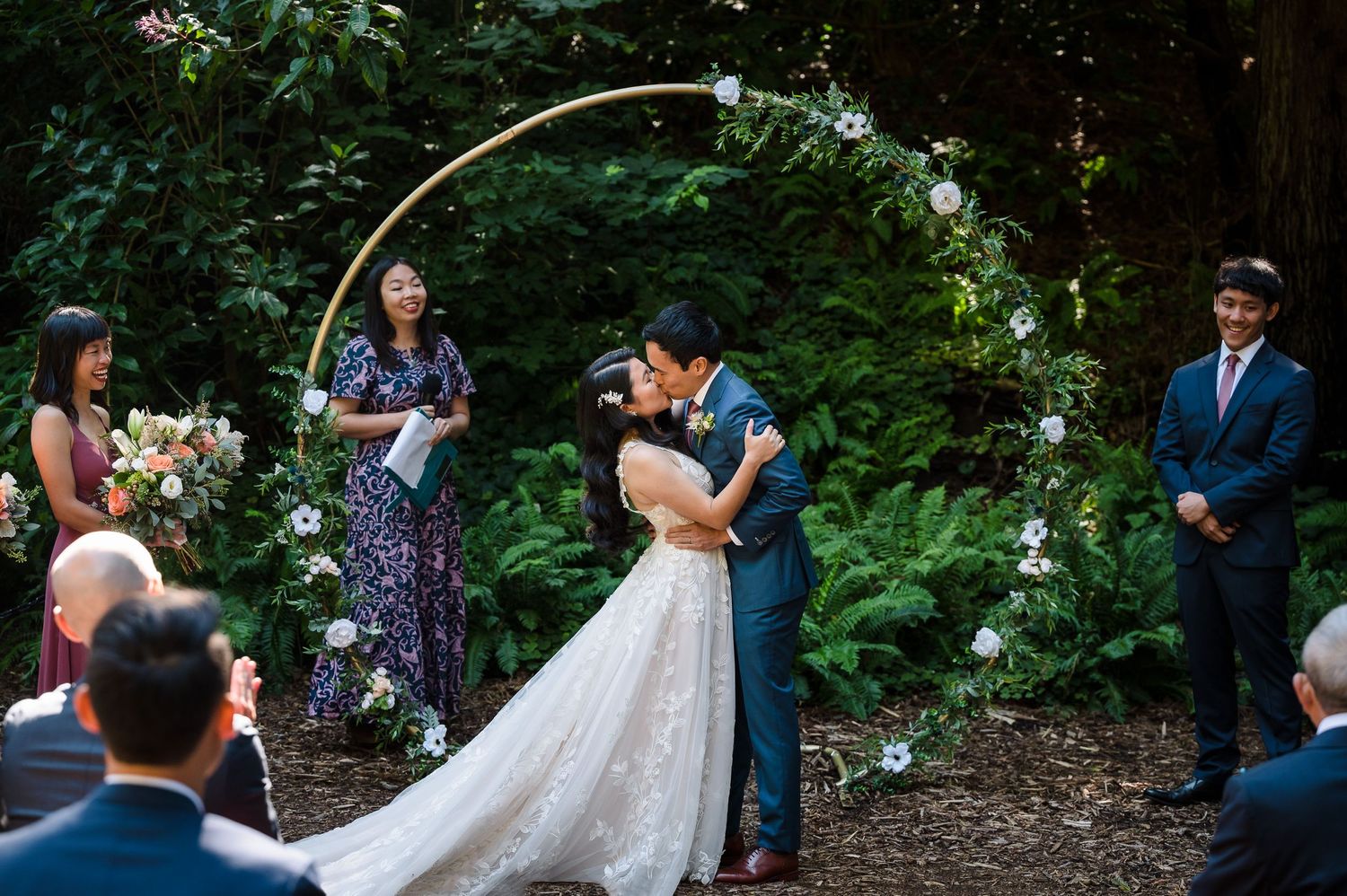 A wedding ceremony moment under an elegant white floral arch in a forest setting.