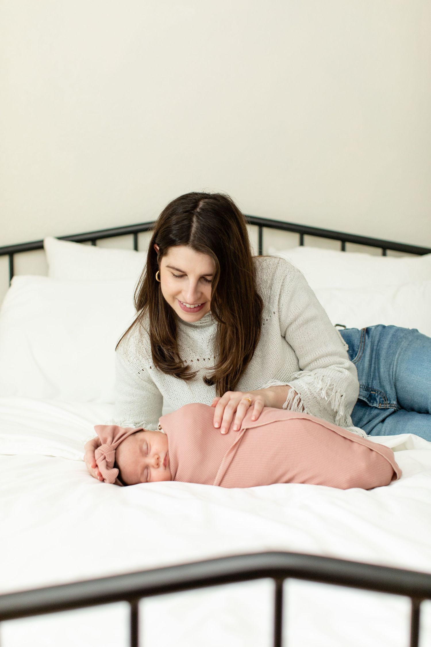 Woman in white sweater lying on bed next to sleeping baby wrapped in pink swaddle.