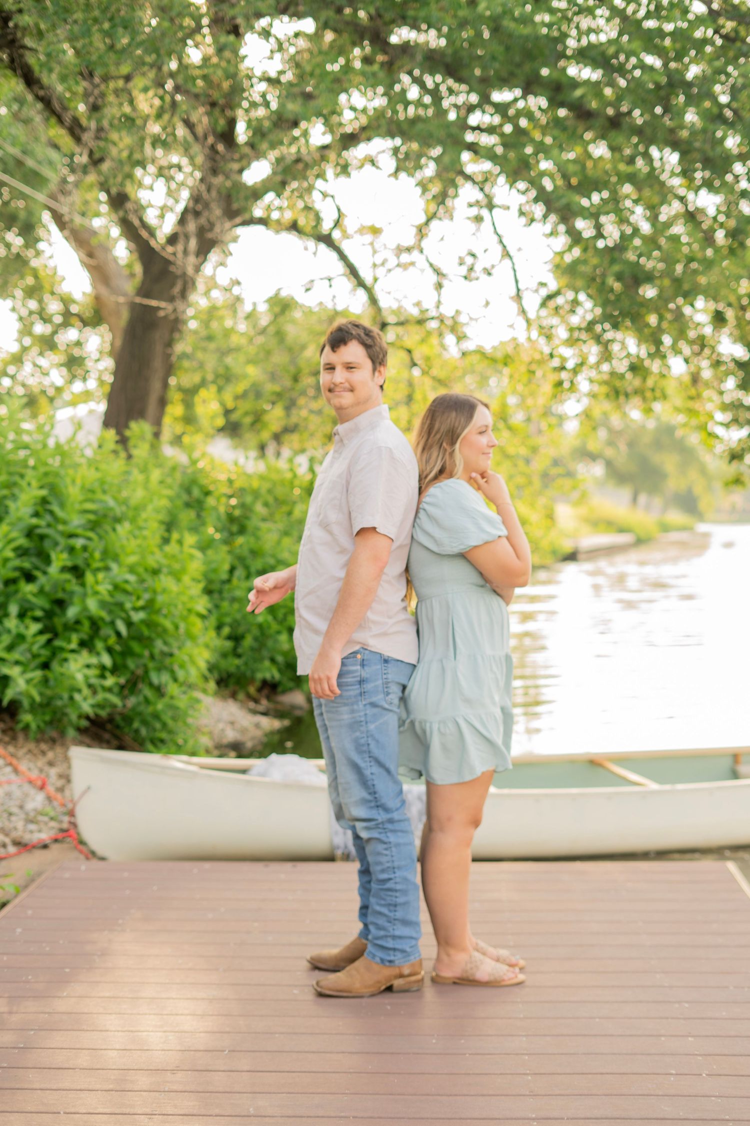 Couple posing back-to-back on a lakeside dock with a white canoe and lush greenery.