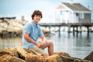 A person sits on coastal rocks with a stilted beach house in the background.