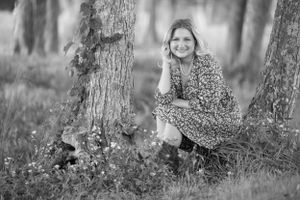 Black and white image of a person crouching by trees in a floral dress.