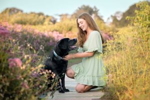 Person in mint green dress kneeling beside black dog on wooden path surrounded by wildflowers.