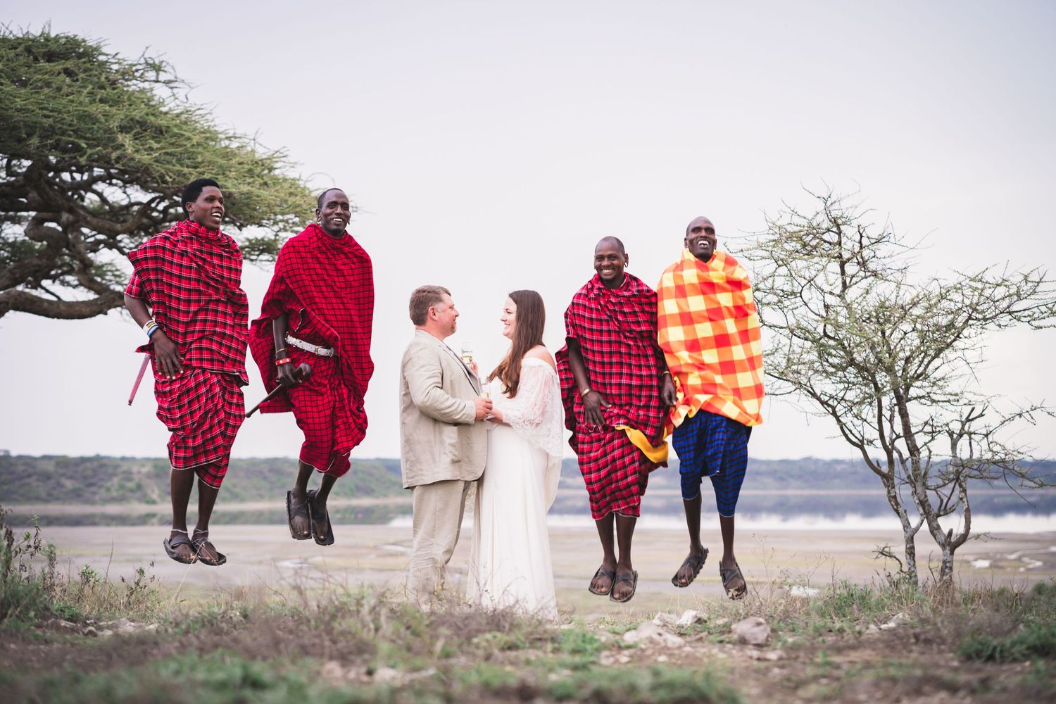 A bride and groom celebrate with jumping Maasai warriors in traditional red shukas outdoors.