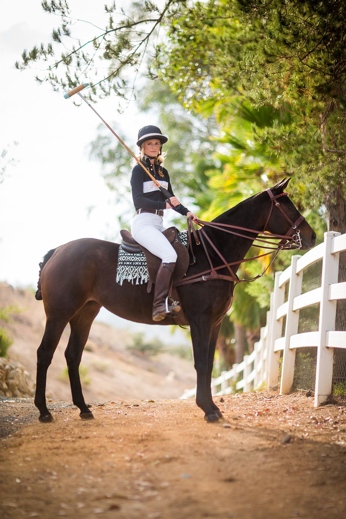 Equestrian in competition attire riding brown horse along white fence trail.