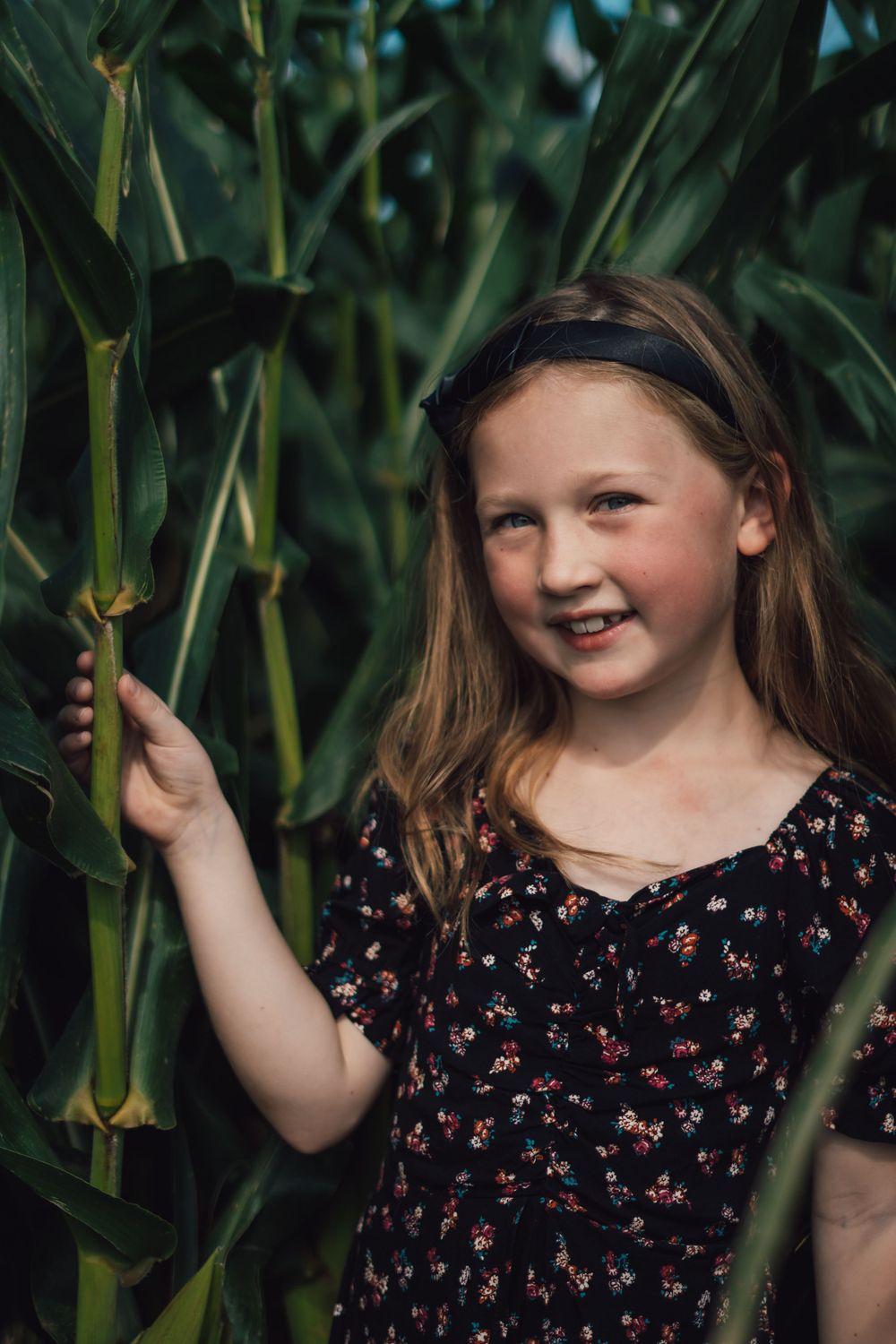 Young girl in floral dress smiling while standing in a cornfield surrounded by tall green stalks.