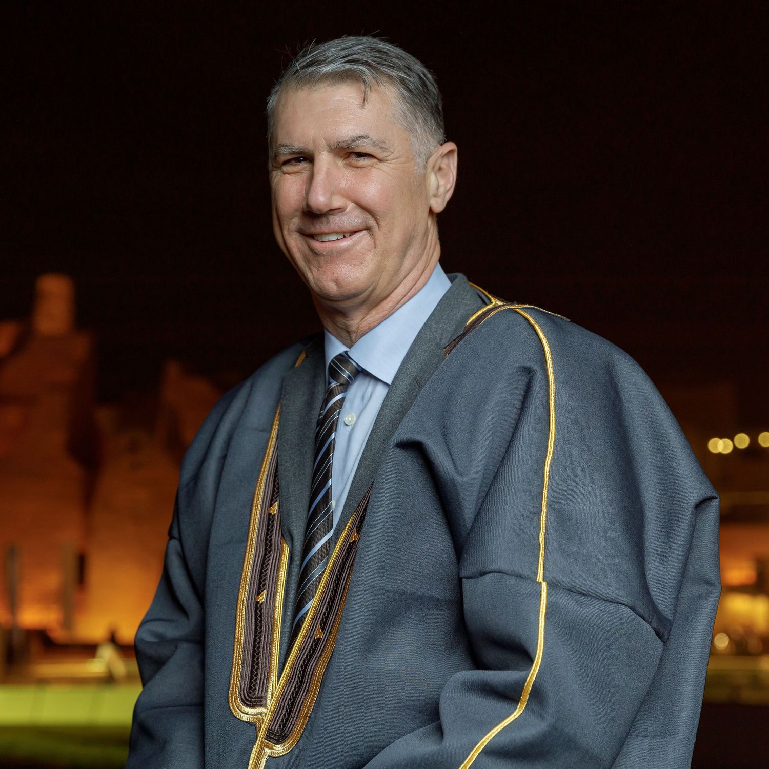 Man in gray suit and tie poses outdoors at night near illuminated buildings.