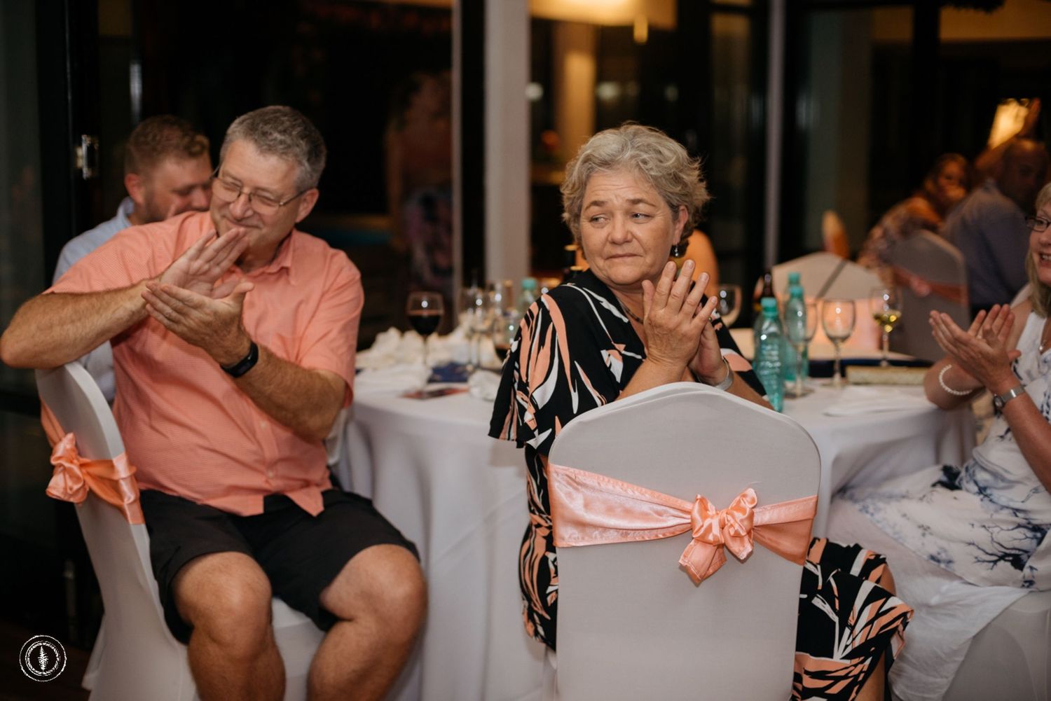 Wedding guests enjoying themselves at a reception with decorated chairs featuring coral-colored bows.
