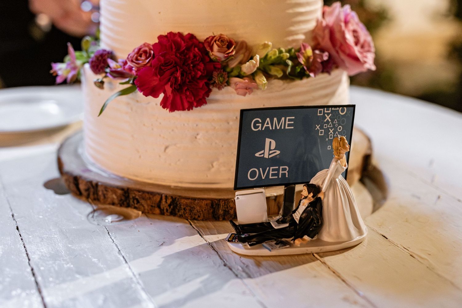 Wedding cake with red flowers decorated with a PlayStation-themed 'Game Over' sign and cake topper on a rustic wood slice.