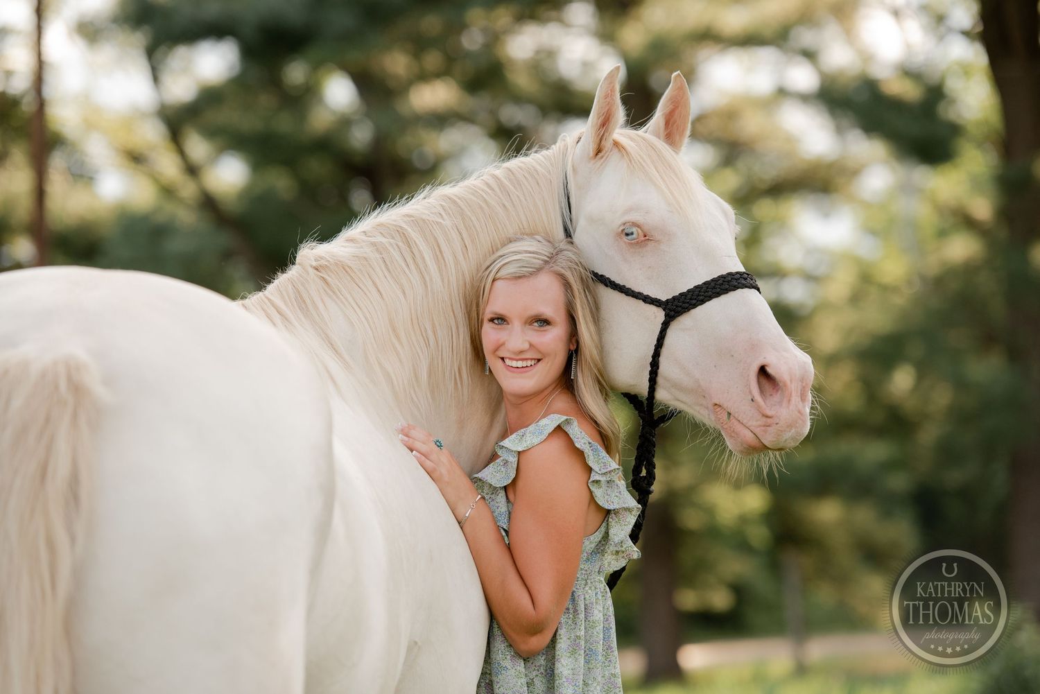Beautiful white horse with black halter poses with rider in sundress against green summer background.