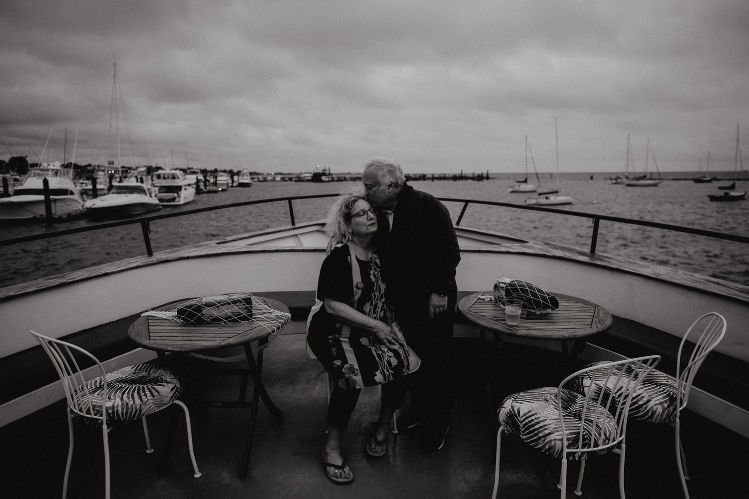 A couple shares an intimate moment on a boat deck overlooking a marina on an overcast day.