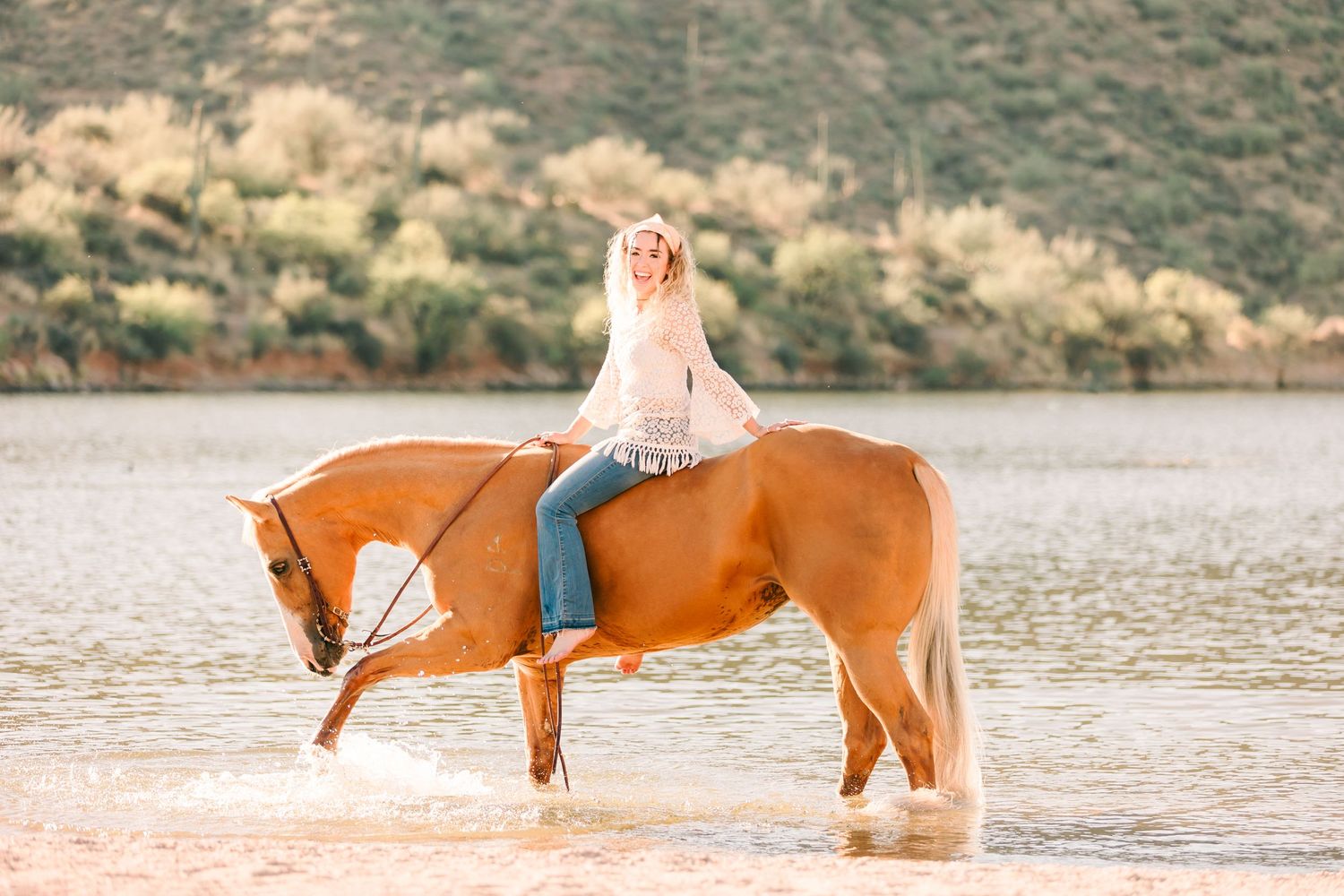 Horseback rider in light clothing atop a brown horse standing in shallow water with mountain backdrop.
