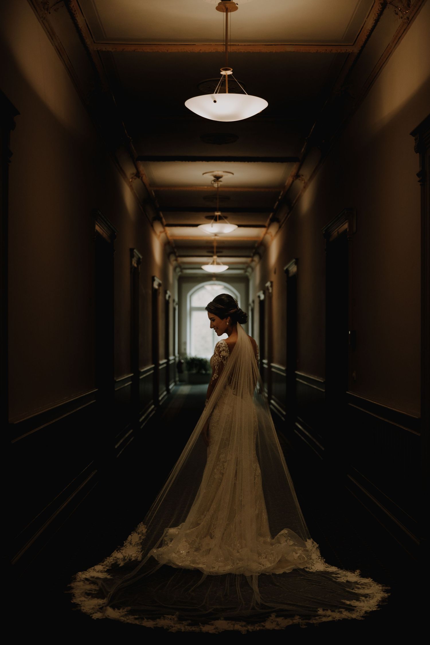 A moody silhouette of a bride in a cathedral veil standing in a dimly lit hallway.