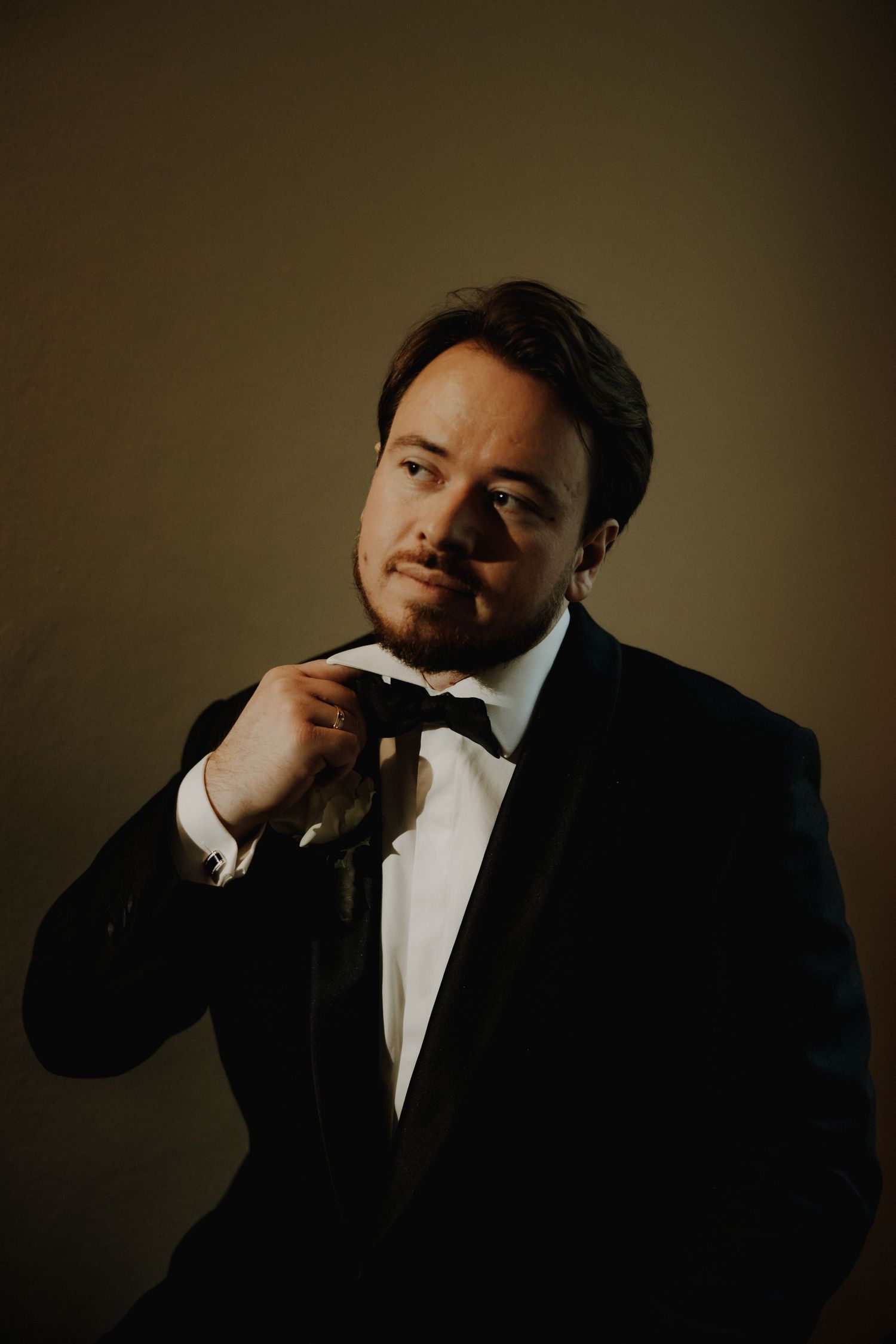 A person in a black tuxedo and bowtie poses against a dark background in a formal portrait.