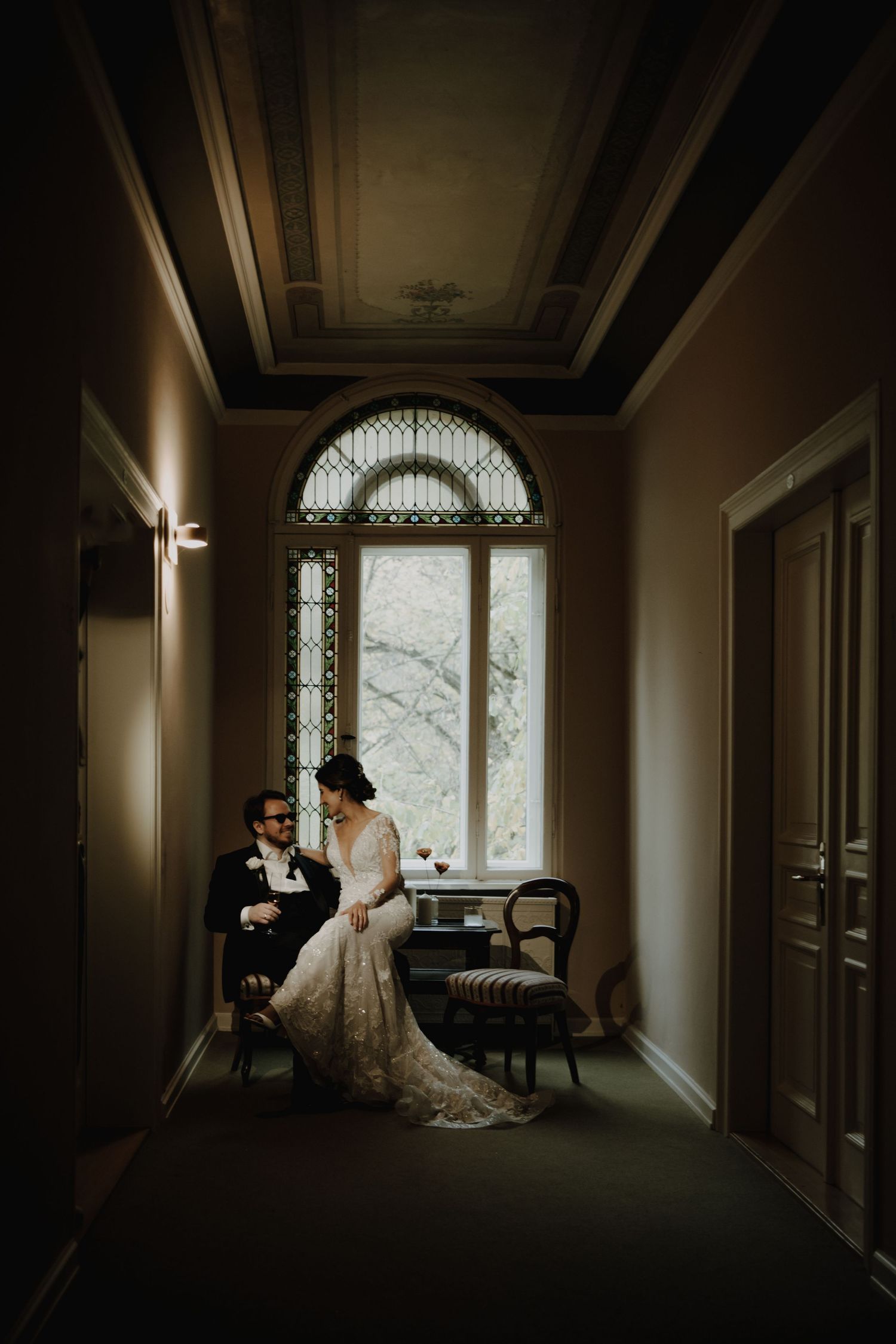 A couple shares an intimate moment in an elegant hallway with arched window and dramatic lighting.