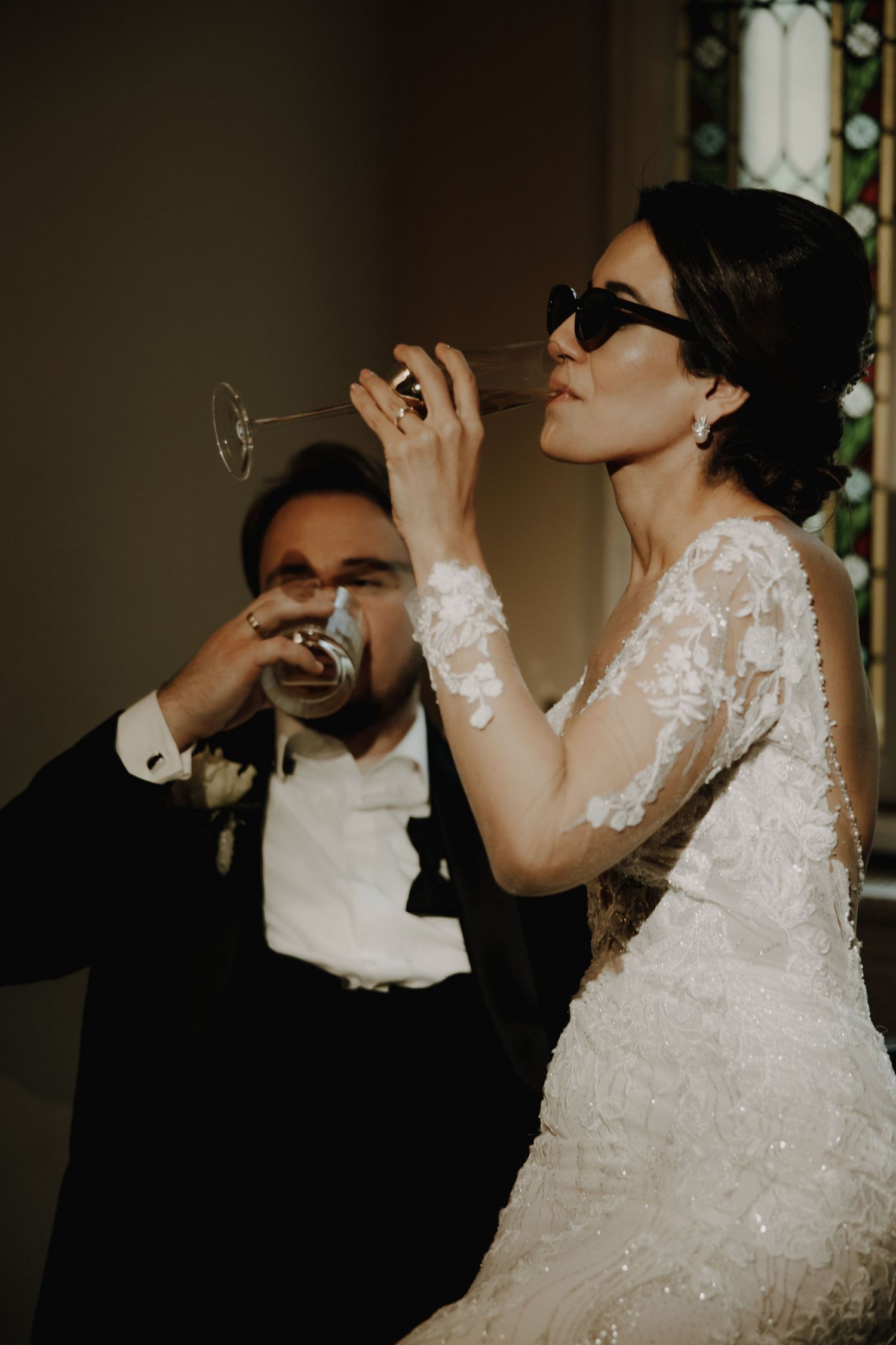 A couple in wedding attire playfully drink from champagne glasses wearing sunglasses.