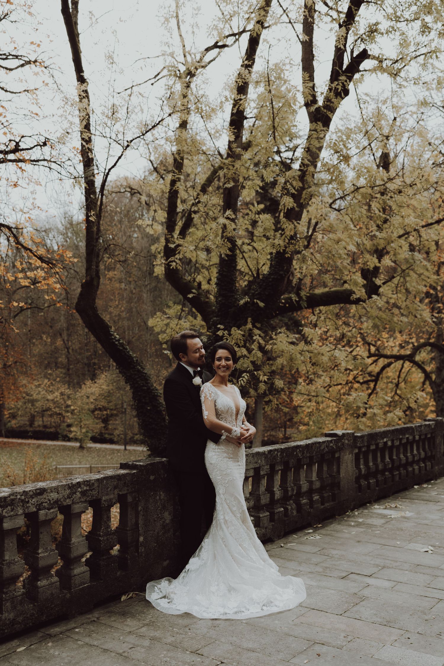 A romantic autumn wedding portrait of a couple standing along a stone balustrade surrounded by fall foliage.