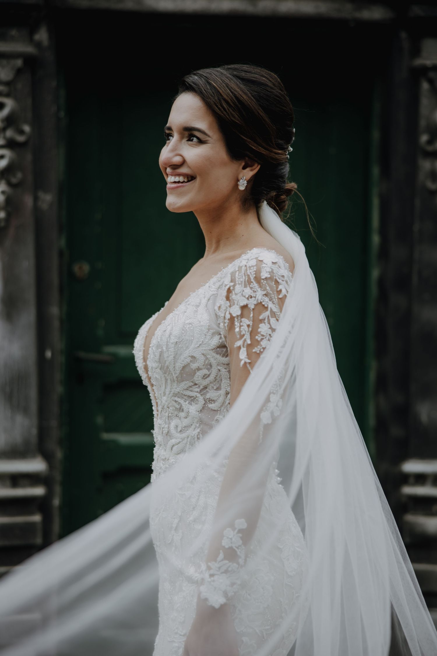 A bride wearing an elegant lace wedding gown with sheer sleeves smiles while standing next to a green door.