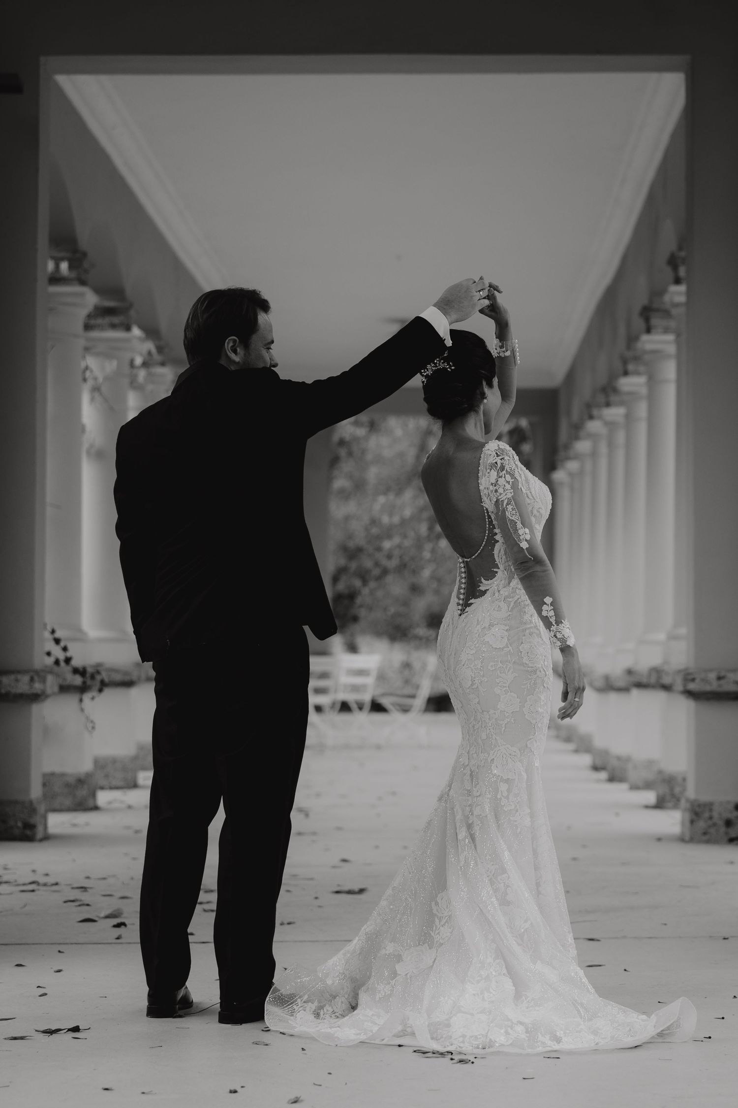 A black and white wedding photo of a couple standing under a columned portico with a lace dress train.