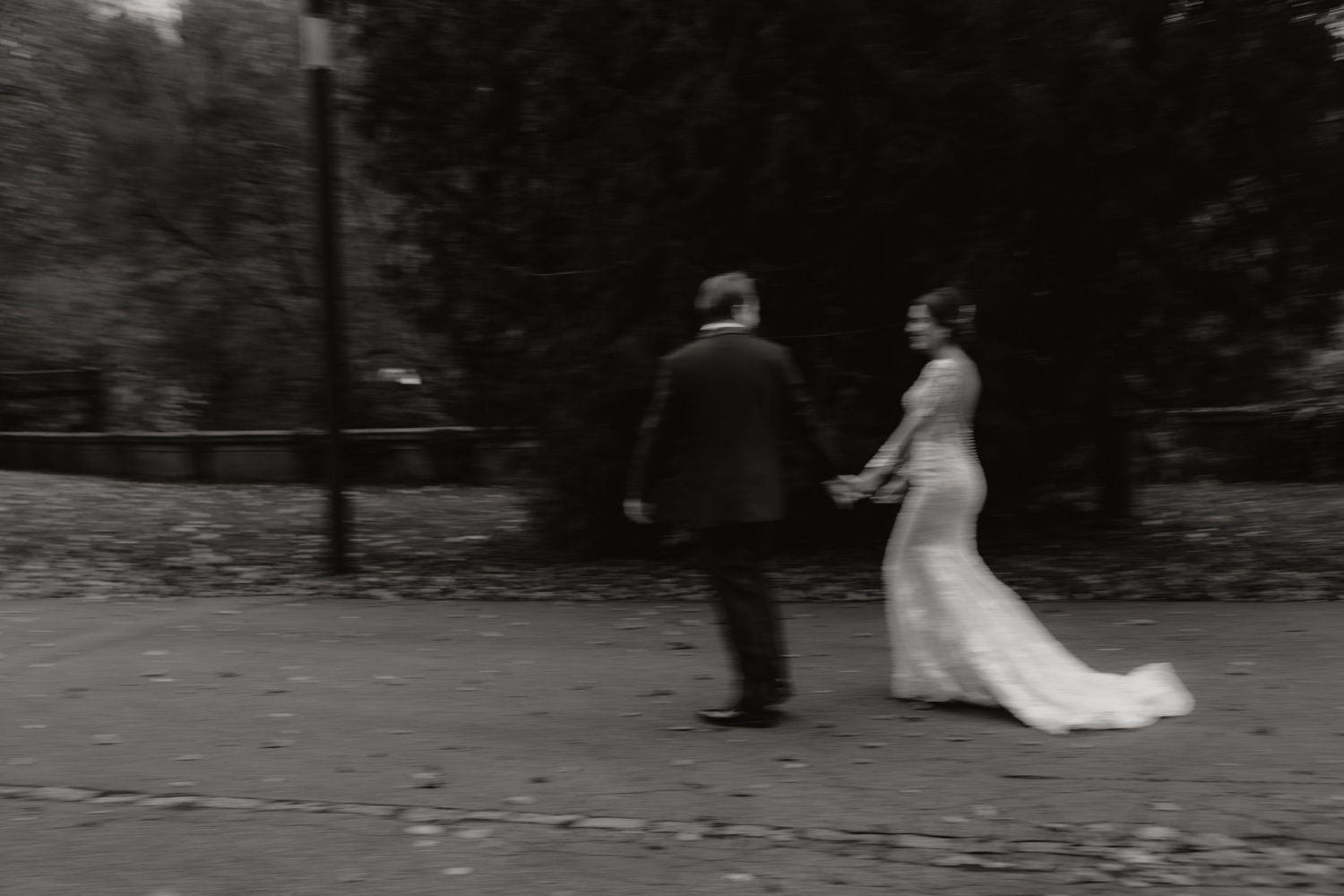Black and white photo of newlyweds walking through a park with trailing wedding dress.