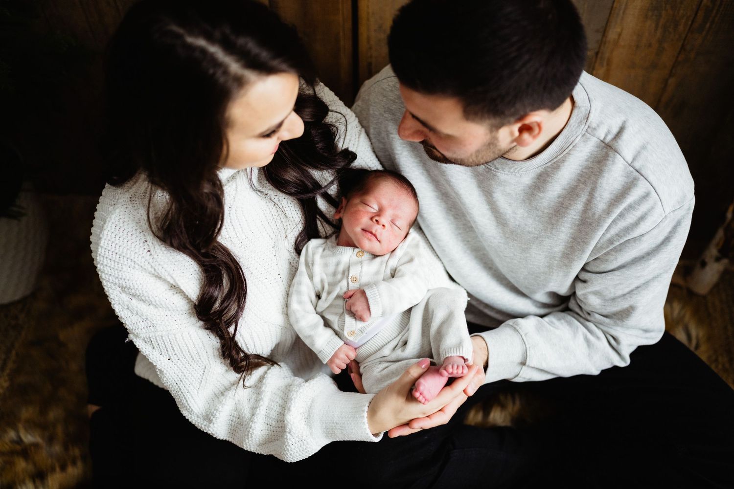 Parents in matching white sweaters gazing lovingly at their newborn baby.