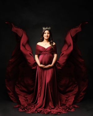 Dramatic maternity portrait with flowing burgundy dress and crown against black background.