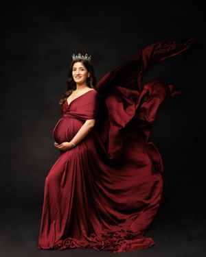Dramatic maternity portrait with flowing burgundy dress and crown against dark backdrop.